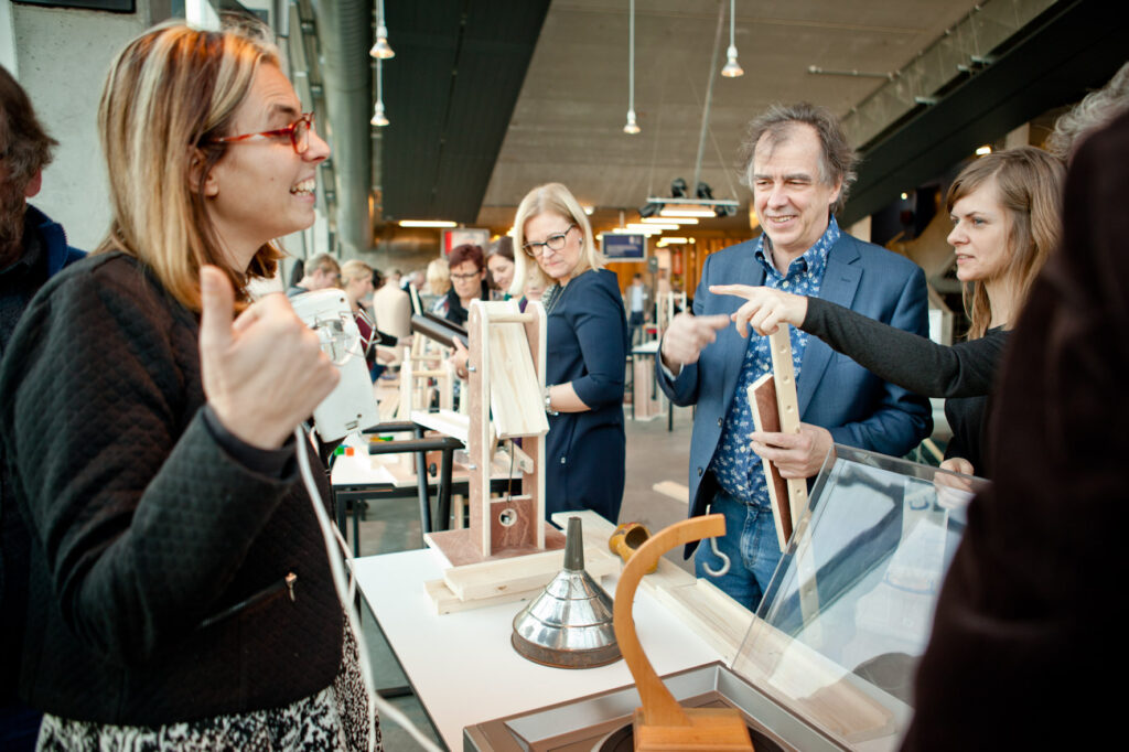 Menschen bei einem Teamevent mit Kettenreaktion sprechen begeistert über technische Bauelemente an einem Indoor-Workshop-Tisch.